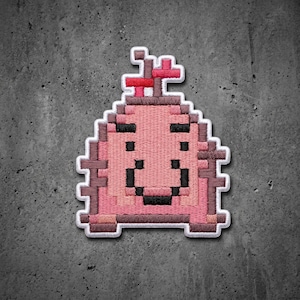 May include: A pixel art style embroidered patch featuring a pink, dome-shaped creature with a dark brown outline. The design includes black pixelated eyes and a mouth, with a small red and brown structure on top, all set against a white border.