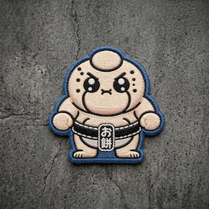 May include: An embroidered patch featuring a cartoon sumo wrestler character with a beige complexion, large black eyes, and a blue border. The character wears a black belt with a white rectangular center containing the Japanese text お餅.