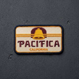 May include: A rectangular embroidered patch with a cream-colored background and a dark blue border. The design features a yellow bell icon above the dark red text PACIFICA and the smaller yellow text CALIFORNIA, all centered on the patch.