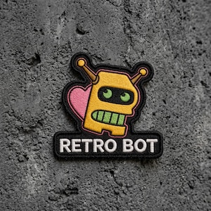 May include: An embroidered patch featuring a yellow robot head with two antennae, green eyes, and a green mouth. A pink heart is visible behind the robot head. The black base of the patch displays the white text RETRO BOT in capital letters.