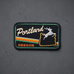 May include: A rectangular embroidered patch with a black background. It features the white text Portland, an orange and green striped line, a white leaping deer silhouette, a yellow outline of the state of Oregon, and the orange text OREGON.