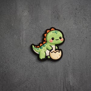 May include: An embroidered patch featuring a small, light green dinosaur with dark green spots, orange back spikes, and pink cheeks. The dinosaur is standing next to a broken, cream-colored eggshell with a black border outlining the entire shape.