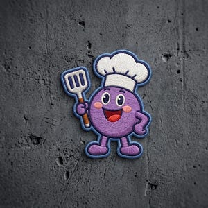May include: An embroidered patch featuring a round purple character with a smiling face, pink cheeks, and a white chef hat. The character holds a white spatula with blue details in one hand and has a blue border outlining the entire shape.