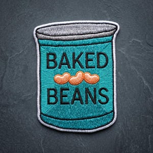 May include: An embroidered patch shaped like a tin can with a teal background, a grey lid, and three orange beans in the center. The text BAKED BEANS is stitched in black capital letters above and below the beans, all outlined in white thread.