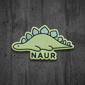 May include: An embroidered patch featuring a light green dinosaur with a row of pale blue plates along its back. The dinosaur is depicted in a side profile view with a simple black outline. Below the dinosaur, the word NAUR is stitched in black.