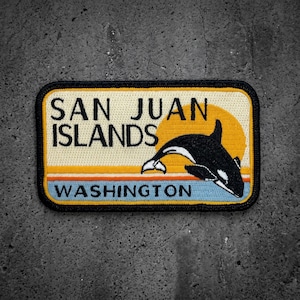 May include: A rectangular embroidered patch featuring a black and white orca whale leaping in front of a yellow sun. The text reads SAN JUAN ISLANDS and WASHINGTON. The design includes horizontal stripes in orange, white, and light blue colors.