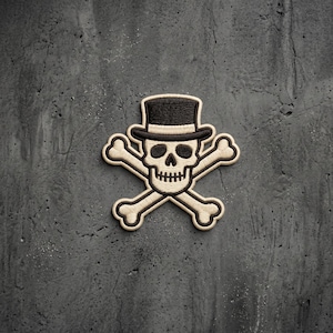 May include: An embroidered patch featuring a skull wearing a black top hat, positioned above two crossed bones. The design uses cream-colored thread for the skull and bones, with black thread for the hat and facial details, creating a high-contrast look.