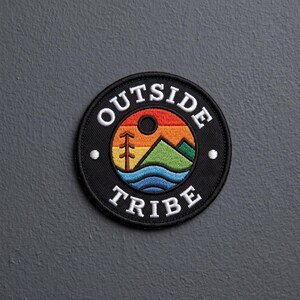 May include: A circular embroidered patch with a black border and the white text OUTSIDE TRIBE. The center features a landscape graphic with a red, orange, and yellow sky, a brown tree, green mountains, and blue water waves in a circular frame.