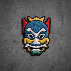 May include: An embroidered patch featuring a stylized mask design with a blue face, white accents around the eyes and nose, red horns, a yellow crown and cheek details, and a green chin section, all outlined in black thread on a textured background.
