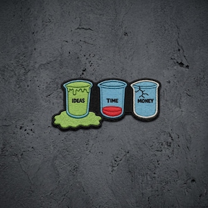 May include: An embroidered patch featuring three cups. The first cup is green with the word IDEAS and a green spill. The second cup is blue with the word TIME and a red base. The third cup is blue with the word MONEY and a crack on the rim.