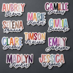 May include: A collection of nine embroidered name patches featuring various first names in colorful, block-style lettering with a cursive surname underneath. Each patch has a white border and displays names such as Aubrey Brown, Camille Dubois, Selena Romano, Marie Fischer, Giulia Bianchi, Claire Dubois, Timson Moreau, Emily Johnson, Madlyn Fischer, and Jessica Wagner.