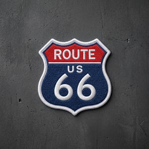 May include: A shield-shaped embroidered patch featuring the text ROUTE US 66. The top section is red with white text, while the larger bottom section is dark blue with white text and numbers. The entire patch has a thick white stitched border.