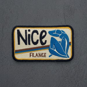 May include: A rectangular embroidered patch with a cream background, a dark blue border, and yellow trim. It features the black text Nice and brown text FRANCE, with a blue stylized figure of a seated person and horizontal brown and blue stripes.