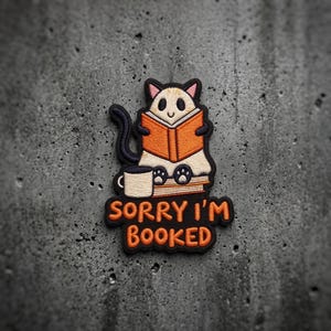 May include: An embroidered patch featuring a cream-colored cat with black outlines reading an orange book while sitting on a stack of books next to a white mug. The text SORRY I'M BOOKED is embroidered in orange letters on a black background.