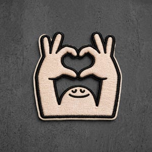 May include: An embroidered patch featuring a beige cartoon character with two hands raised above its head, forming a heart shape with its fingers. The design is outlined in thick black thread, with a simple curved line for a mouth and closed eyes.
