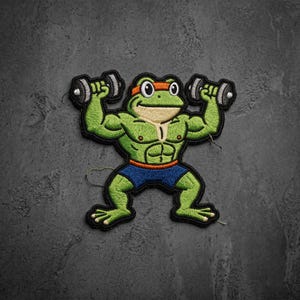 May include: An embroidered patch featuring a muscular green frog wearing blue shorts and an orange headband. The frog is depicted with a tan chest and belly, lifting two grey dumbbells, one in each hand, with a black border outlining the entire shape.