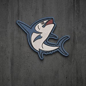 May include: An embroidered patch featuring a stylized tuna fish in profile. The fish has a dark blue outline and fins, a white body with fine stitching detail, and a small red accent inside the open mouth, all set against a dark gray background.