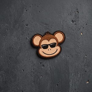 May include: An embroidered patch featuring a cartoon monkey face with brown fur, large rounded ears, and black sunglasses. The design is outlined in a thick black border, showcasing detailed stitching that defines the facial features and expression.