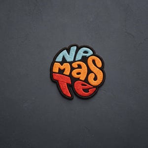 May include: A circular embroidered patch featuring the word NAMASTE in a stylized, stacked font. The letters NA are light blue, MAS are orange, and TE are red. The entire design is outlined in black thread with a thick, dark border.