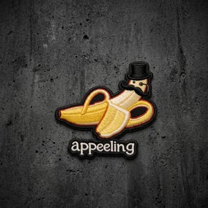 May include: An embroidered patch featuring a yellow banana wearing a black top hat and a black handlebar mustache. Below the banana, the word appeeling is embroidered in white lowercase letters with a black outline on a black background.