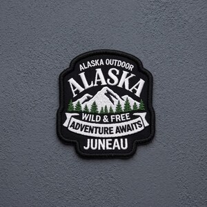 May include: A black embroidered patch featuring white text that reads Alaska Outdoor, Alaska, Wild & Free, Adventure Awaits, and Juneau. The design includes a white mountain range with green pine trees at the base, all enclosed in a black border.