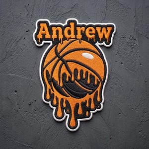May include: An embroidered patch featuring the name Andrew in orange text with a black outline, positioned above a basketball graphic. The basketball design incorporates a dripping effect in orange and black, with a white border surrounding the entire piece.