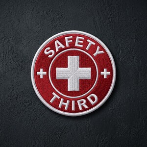 May include: A circular embroidered patch featuring a deep red background with a white border. In the center is a white cross, flanked by two small white plus signs. The text SAFETY is arched above the cross, and THIRD is arched below.