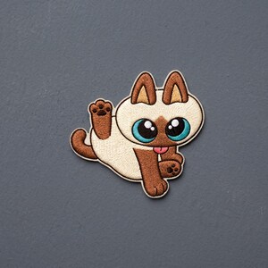 May include: An embroidered patch featuring a cream and brown Siamese-style cat with large, bright blue eyes, pointed ears, and a small pink tongue sticking out. The cat is depicted in a playful pose with one paw raised and a curved tail.