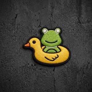 May include: An embroidered patch featuring a bright green frog sitting inside a yellow rubber duck floatie. The frog has white eyes with black pupils, and the duck has a distinct orange beak. The entire design is outlined in black thread.