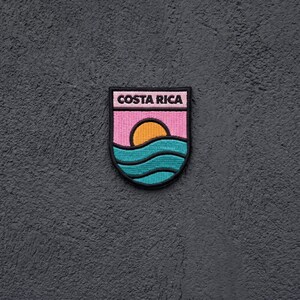 May include: A shield-shaped embroidered patch featuring the text COSTA RICA in black letters at the top. The design includes a yellow sun rising over teal blue ocean waves against a pink sky background, all outlined with a thick black border.