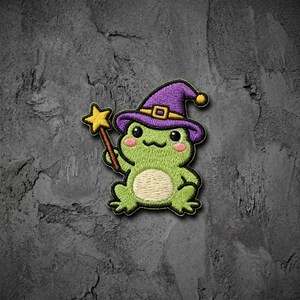May include: An embroidered patch featuring a light green frog wearing a purple wizard hat with a yellow band and buckle. The frog has pink cheeks, a cream-colored belly, and is holding a yellow star-tipped magic wand in its right hand.