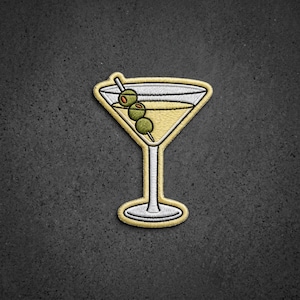 May include: An embroidered patch shaped like a martini glass with a pale yellow liquid inside. Three green olives with small orange centers are skewered on a silver toothpick resting across the rim of the glass. The patch has a thick yellow border.