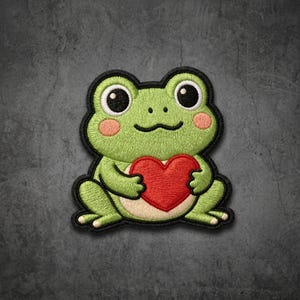 May include: An embroidered patch featuring a light green frog with large black eyes, pink cheeks, and a cream-colored belly. The frog is holding a bright red heart against its chest, with the entire design outlined in a thick black border.