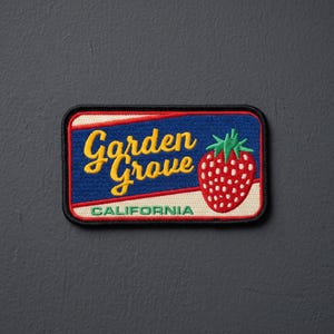 May include: A rectangular embroidered patch featuring a blue background with the yellow text Garden Grove and the green text CALIFORNIA. A red strawberry with green leaves is on the right side, all bordered by a thick black stitched edge.