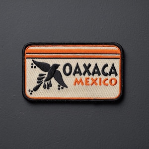 May include: A rectangular embroidered patch with a black border. The design features a cream background with an orange horizontal stripe at the top. It displays a black bird silhouette and the black text OAXACA above the orange text MEXICO.