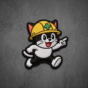 May include: An embroidered patch featuring a black and white cat wearing a yellow construction hard hat with a green cross symbol. The cat is depicted in a running pose with one paw pointing forward, featuring a red tongue and black outlines.