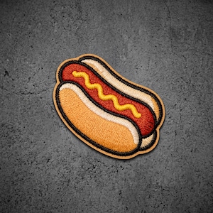 May include: An embroidered patch shaped like a hot dog in a bun. The design features a red sausage nestled inside a tan bun, topped with a wavy line of yellow mustard. The entire motif is outlined in thick black thread, creating a bold appearance.