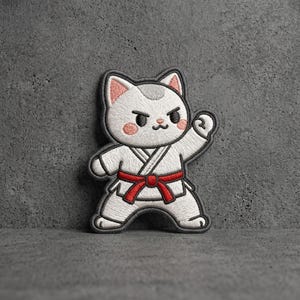 May include: An embroidered patch featuring a white cat with pink inner ears and cheeks, wearing a white martial arts uniform with a red belt. The cat is in a dynamic pose with one fist raised, outlined in dark gray thread against a textured background.