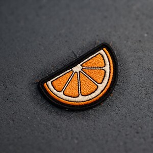 May include: An embroidered patch in the shape of a citrus orange slice. The design features bright orange segments outlined in white thread, all enclosed within a thick, solid black border that defines the semi-circular shape of the fruit slice.