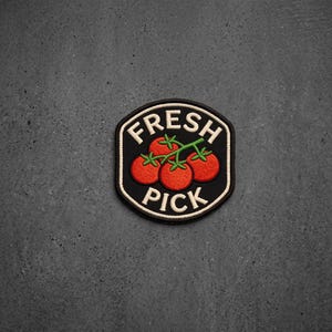 May include: A circular embroidered patch with a black background and a white border. The design features three red tomatoes on a green vine in the center. The text FRESH is embroidered in white above the tomatoes, and PICK is below them.