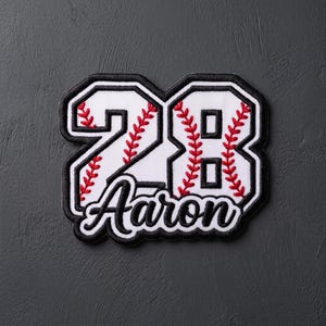 May include: A white embroidered patch featuring the number 28 in a baseball-themed style with red stitching details. The name Aaron is embroidered in black script lettering below the number. The entire design is outlined with a thick black border.
