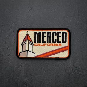 May include: A rectangular embroidered patch with a black border. It features a cream background, a graphic of a tower with a red roof, and the black text "MERCED" above the orange text "CALIFORNIA". A diagonal orange stripe crosses the lower right.