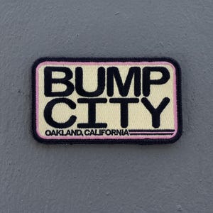 May include: A rectangular embroidered patch with a cream-colored background and a black border. The text BUMP CITY is stitched in large black letters, with OAKLAND, CALIFORNIA in smaller black text below, all framed by a thin pink inner border.