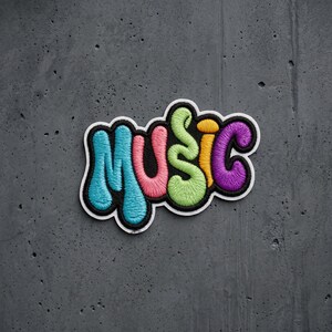 May include: An embroidered patch featuring the word MUSIC in a bubble-style font. Each letter is a different color: the M is light blue, the U is pink, the S is light green, the I is yellow, and the C is purple, all outlined in black and white.