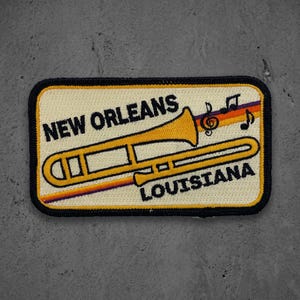 May include: A rectangular embroidered patch with a cream background and a black border. It features a yellow trombone, black text reading NEW ORLEANS and LOUISIANA, and a multi-colored striped line with black musical notes above the instrument.