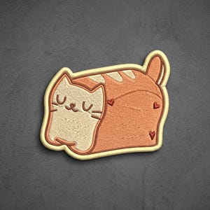 May include: An embroidered patch shaped like a loaf of bread with a cat face on one end. The cat is light cream with closed eyes and whiskers. The body of the bread is orange with darker orange stripes and three small red heart-shaped accents.