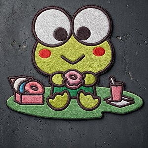 May include: An embroidered patch featuring a green frog character with large white eyes, red cheeks, and a dark brown outline. The frog sits on a green lily pad, holding a pink donut, with a box of donuts and a pink drink with a straw nearby.
