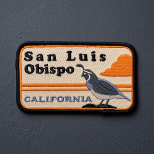 May include: A rectangular embroidered patch with a cream background, black border, and orange horizontal stripes. It features the black text San Luis Obispo, a grey and white California quail illustration, and the blue text CALIFORNIA.