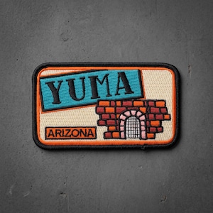May include: A rectangular embroidered patch with a black border. It features a light beige background, a bright blue banner with the black text YUMA, and a red brick wall graphic with a white arched gate. The word ARIZONA is printed in black.