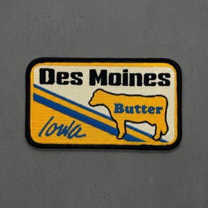 May include: A rectangular embroidered patch with a black border. The background is cream with a yellow section featuring a diagonal blue stripe. It displays the black text Des Moines, the blue text Iowa, and a yellow cow silhouette with the blue text Butter.
